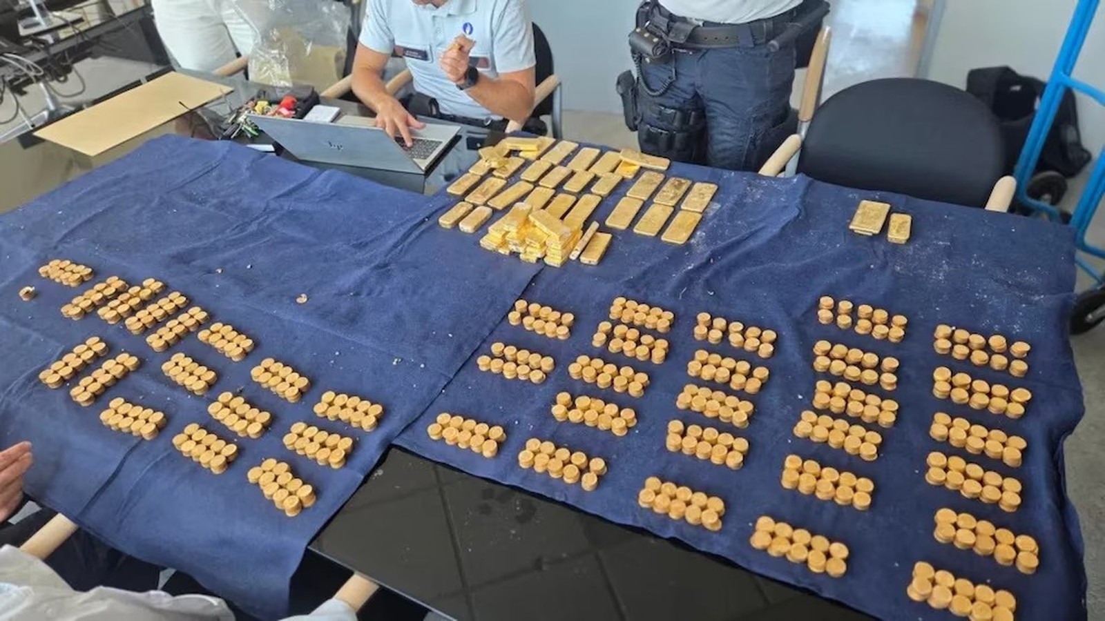 Mesa com pano azul, mostrando barras e pilhas de moedas de ouro, com oficiais