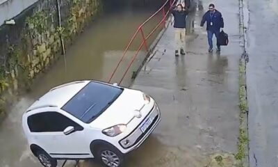 Carro branco parcialmente submerso em rio barrento, dois homens em calçada molhada, um com mãos na cabeça