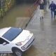 Carro branco parcialmente submerso em rio barrento, dois homens em calçada molhada, um com mãos na cabeça