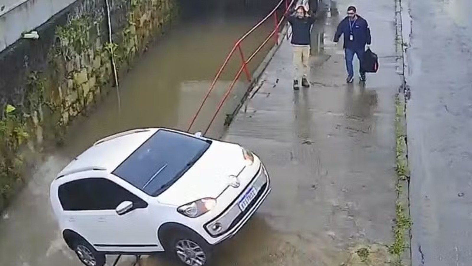 Carro branco parcialmente submerso em rio barrento, dois homens em calçada molhada, um com mãos na cabeça