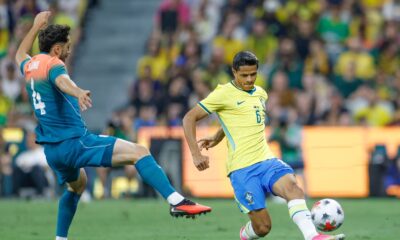 Dois jogadores, um do Brasil e outro adversário (VIANO), disputam a bola em campo de futebol