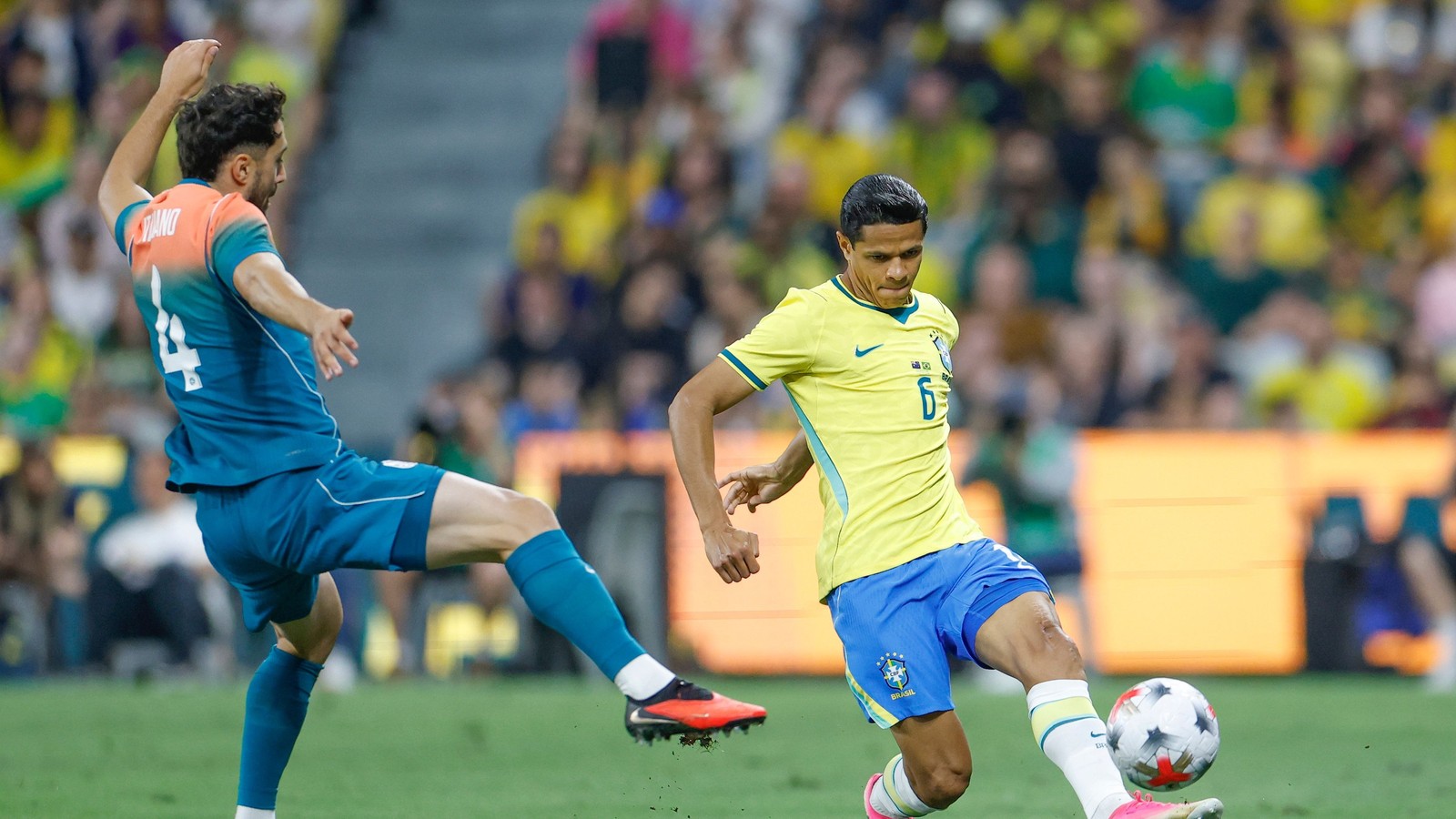 Dois jogadores, um do Brasil e outro adversário (VIANO), disputam a bola em campo de futebol