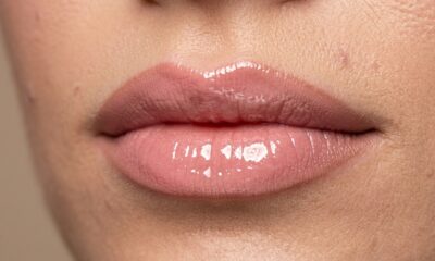 Lábios finos com contorno nude esfumado, batom rosado e gloss aplicado no centro para criar efeito de volume.