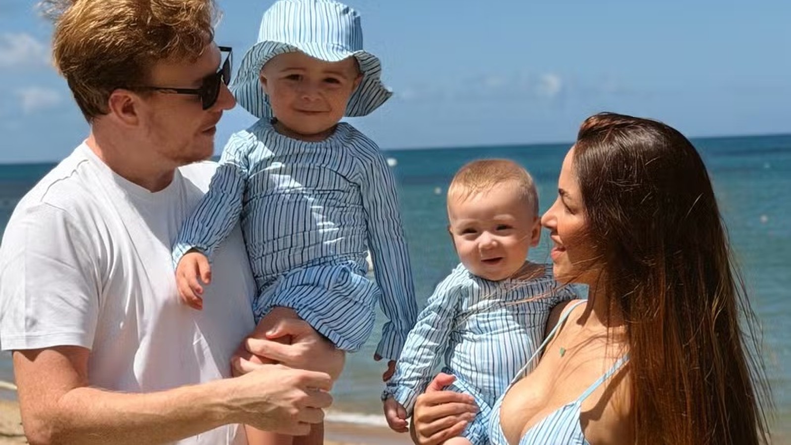 Homem, mulher e dois bebês na praia, todos sorrindo e usando roupas listradas
