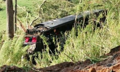 Ônibus escuro danificado, virado em ribanceira com vegetação e terra