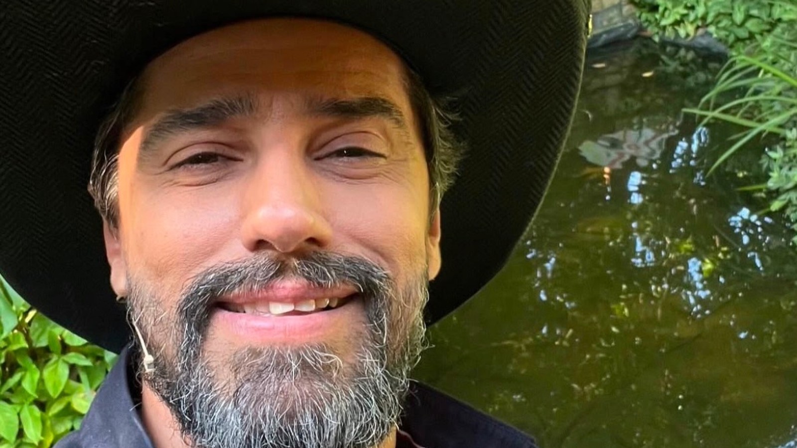 Close-up de homem barbudo grisalho, chapéu escuro, sorrindo, com fundo verde e água