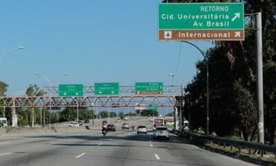 Rodovia com tráfego de veículos, grandes placas de sinalização verdes e pórticos metálicos