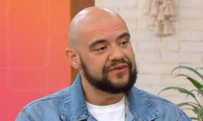 Homem calvo com barba e jaqueta jeans, camiseta branca, falando em estúdio