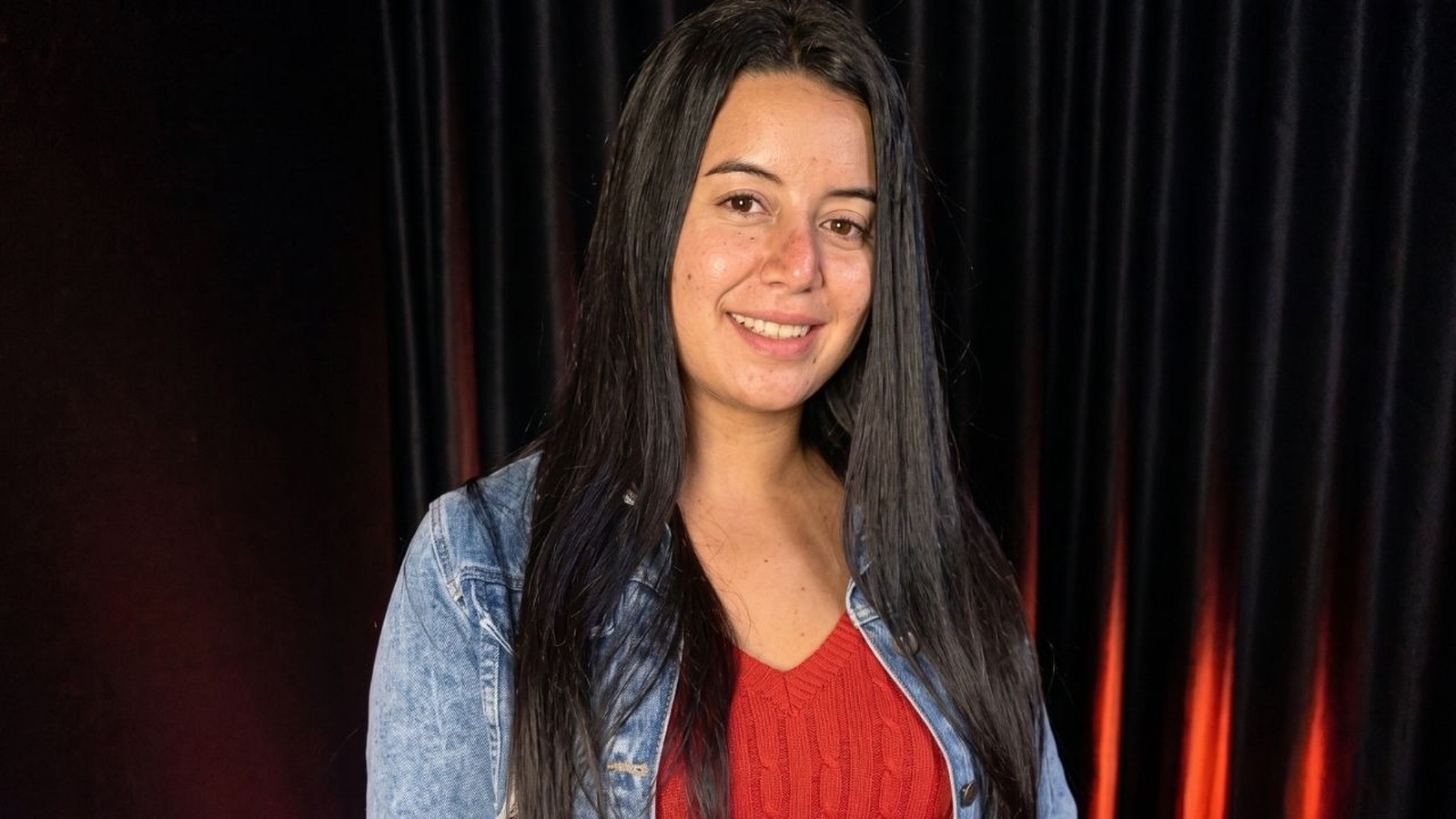 Mulher jovem com cabelos escuros e jaqueta jeans sorri para a câmera