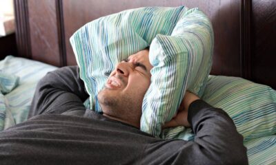 Homem deitado na cama, rosto contorcido de incômodo, cobrindo os ouvidos com travesseiro