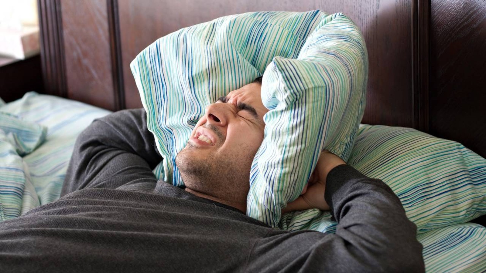 Homem deitado na cama, rosto contorcido de incômodo, cobrindo os ouvidos com travesseiro