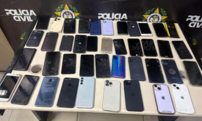 Muitos celulares de diversas marcas e modelos, alguns com telas quebradas, sobre uma mesa policial