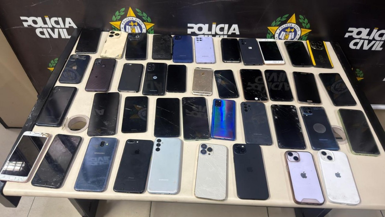 Muitos celulares de diversas marcas e modelos, alguns com telas quebradas, sobre uma mesa policial