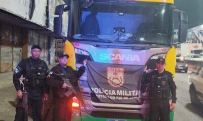 Três policiais fardados com armas e bandeira PMERJ em frente a um caminhão Scania R150
