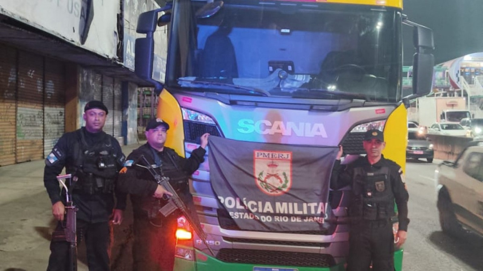 Três policiais fardados com armas e bandeira PMERJ em frente a um caminhão Scania R150
