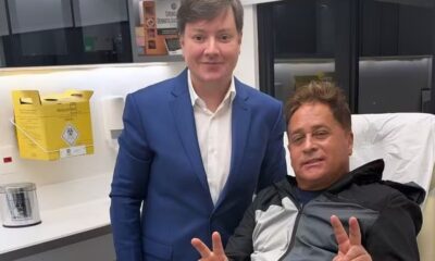 Dois homens sorrindo em clínica. Um de terno, outro de jaqueta faz paz. Lixeira médica
