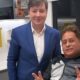 Dois homens sorrindo em clínica. Um de terno, outro de jaqueta faz paz. Lixeira médica