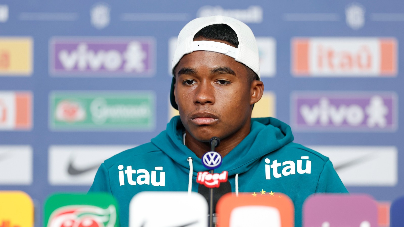 Jovem jogador Endrick em coletiva de imprensa, usando boné branco e moletom teal com logos de patrocinadores