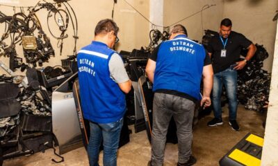 Três homens em galpão cheio de peças automotivas. Dois usam coletes azuis "DETRAN DESMONTE"