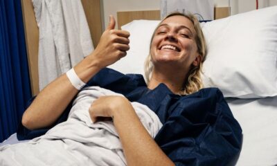 Mulher loira sorri na cama de hospital, olhos fechados, fazendo sinal de positivo