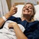 Mulher loira sorri na cama de hospital, olhos fechados, fazendo sinal de positivo