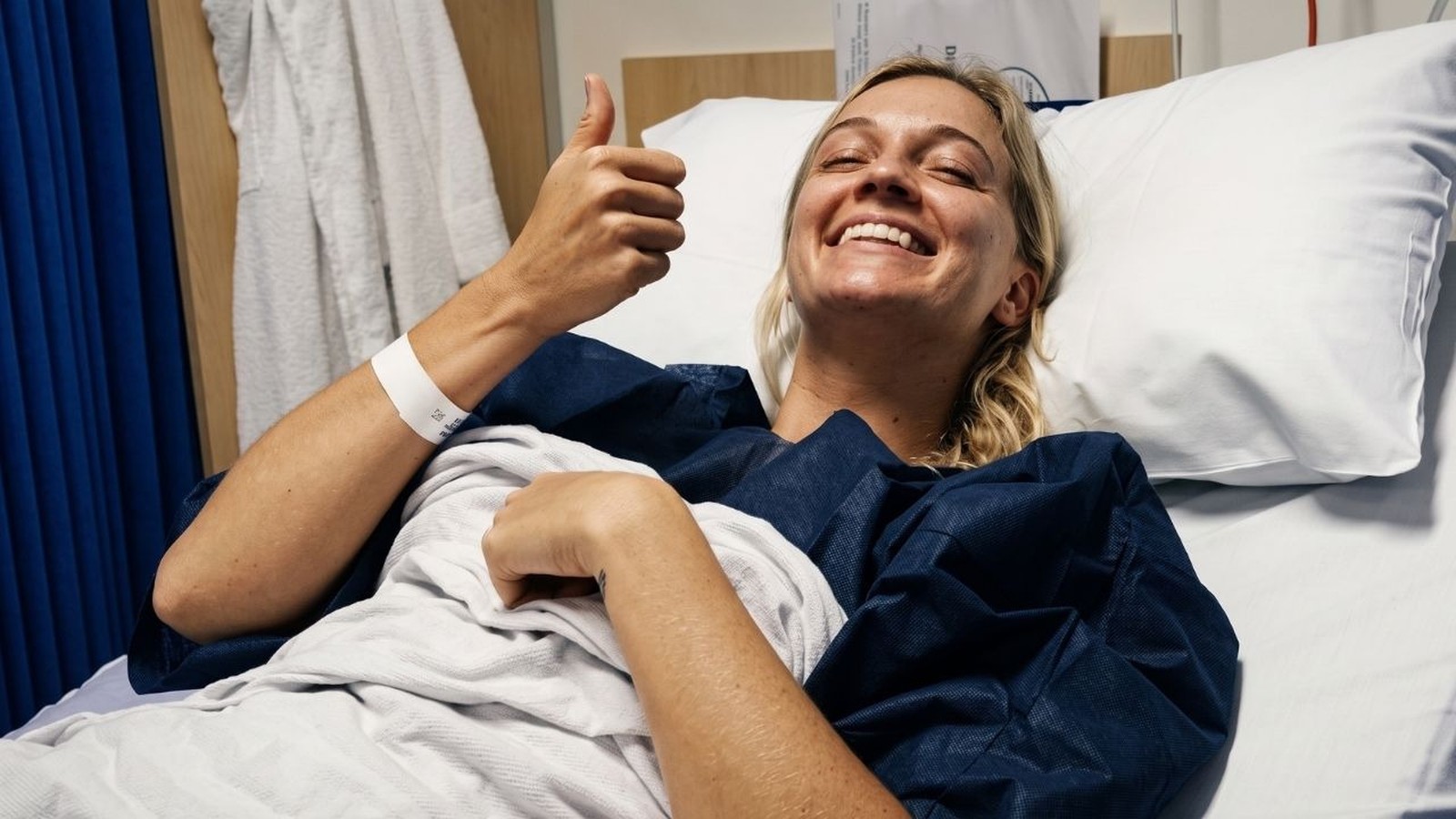 Mulher loira sorri na cama de hospital, olhos fechados, fazendo sinal de positivo
