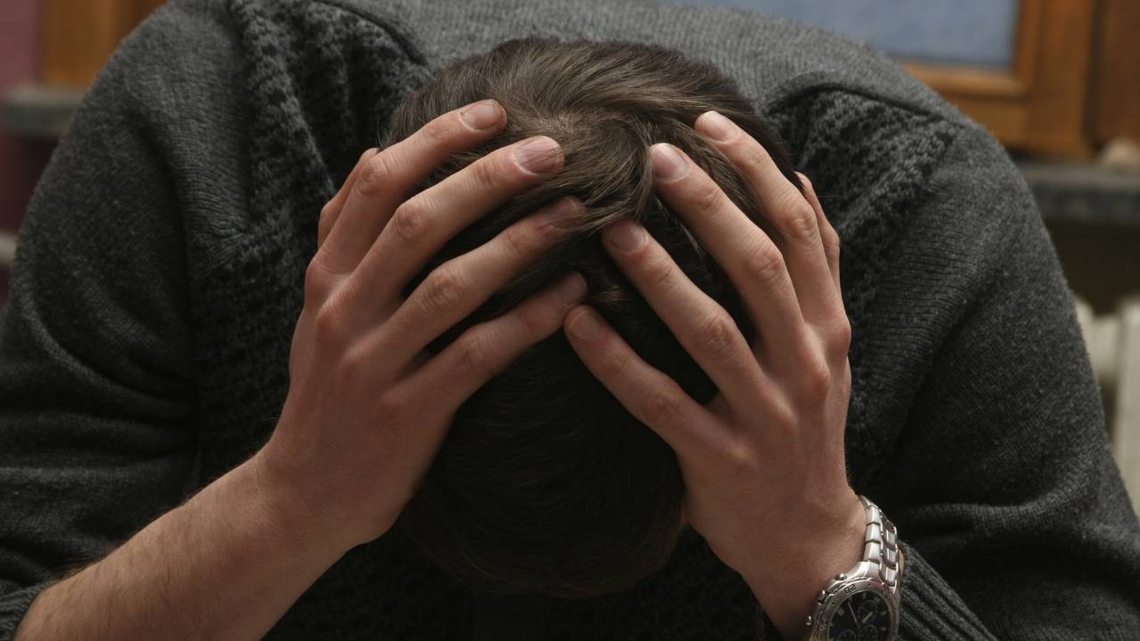 Homem com as mãos na cabeça e a cabeça baixa, vestindo um suéter cinza e relógio