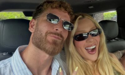 Selfie de casal em carro, mulher loira e homem barbudo sorrindo com óculos escuros