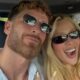 Selfie de casal em carro, mulher loira e homem barbudo sorrindo com óculos escuros