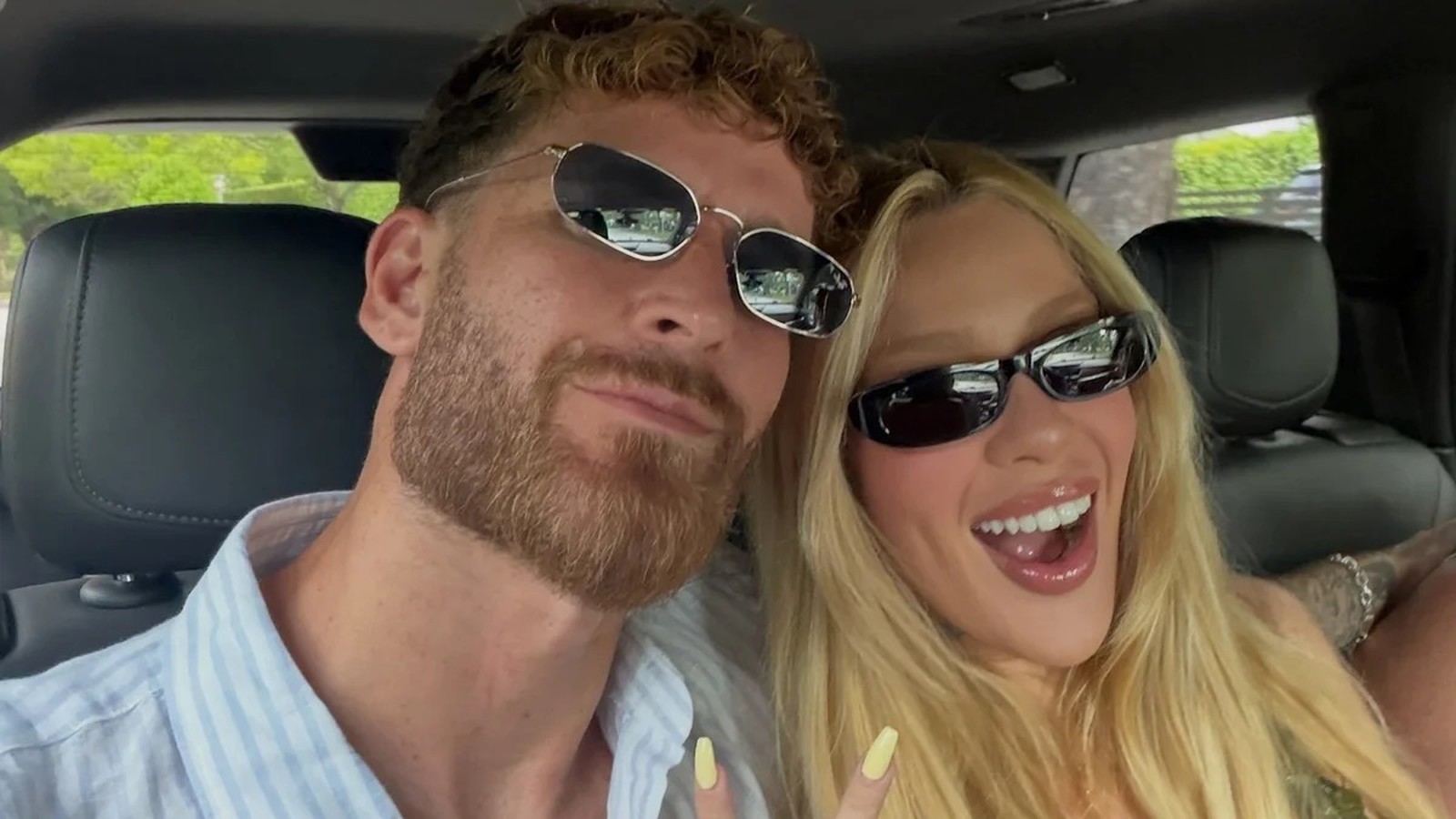 Selfie de casal em carro, mulher loira e homem barbudo sorrindo com óculos escuros