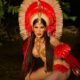 Mulher com cocar de penas vermelhas e douradas, maquiagem facial tribal, agachada em água escura