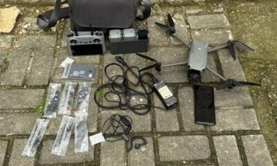 Drone cinza, controle remoto, baterias, hélices lacradas, cabos e celular preto em piso de concreto