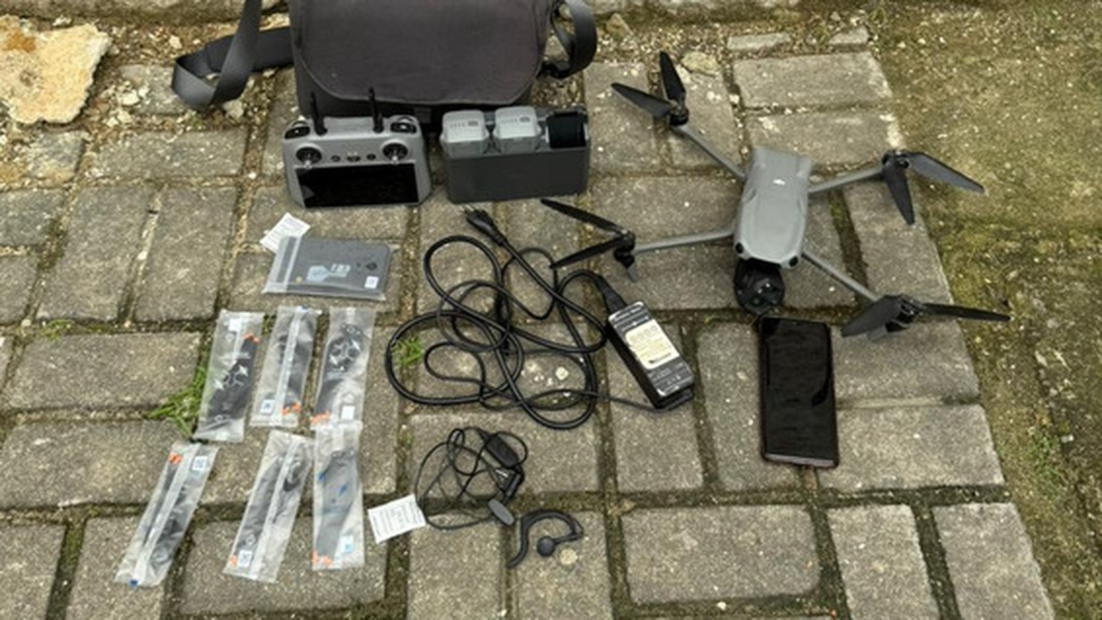 Drone cinza, controle remoto, baterias, hélices lacradas, cabos e celular preto em piso de concreto