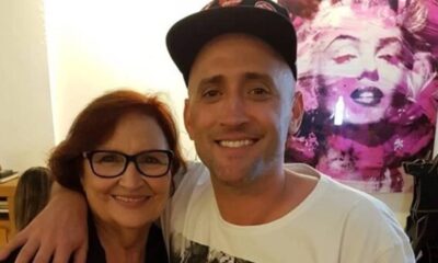 Dona Déa e Paulo Gustavo sorriem abraçados. Mulher de óculos, homem de boné. Quadro ao fundo