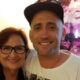 Dona Déa e Paulo Gustavo sorriem abraçados. Mulher de óculos, homem de boné. Quadro ao fundo