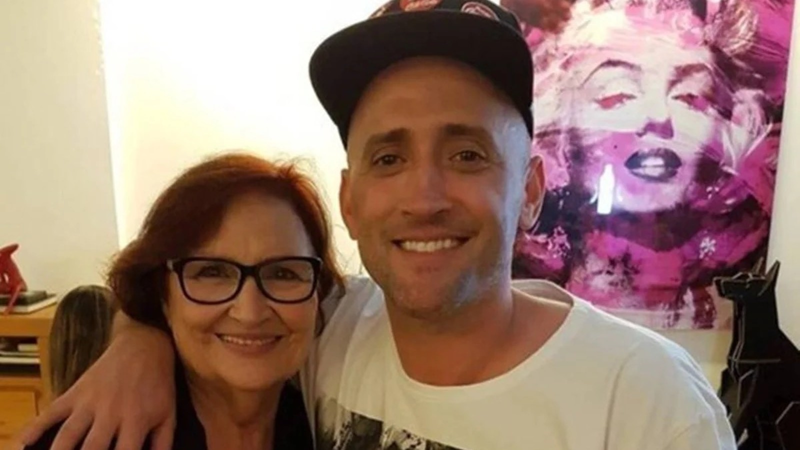 Dona Déa e Paulo Gustavo sorriem abraçados. Mulher de óculos, homem de boné. Quadro ao fundo