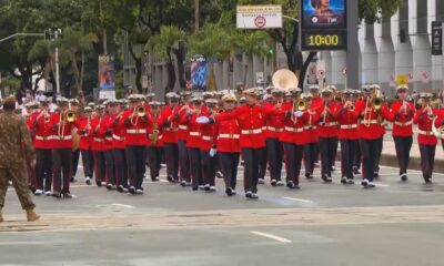 Banda militar em uniforme vermelho desfila tocando instrumentos de sopro em avenida urbana