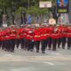 Banda militar em uniforme vermelho desfila tocando instrumentos de sopro em avenida urbana