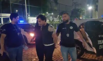 Dois policiais conduzem um homem próximo a uma viatura da Polícia Civil à noite