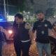 Dois policiais conduzem um homem próximo a uma viatura da Polícia Civil à noite
