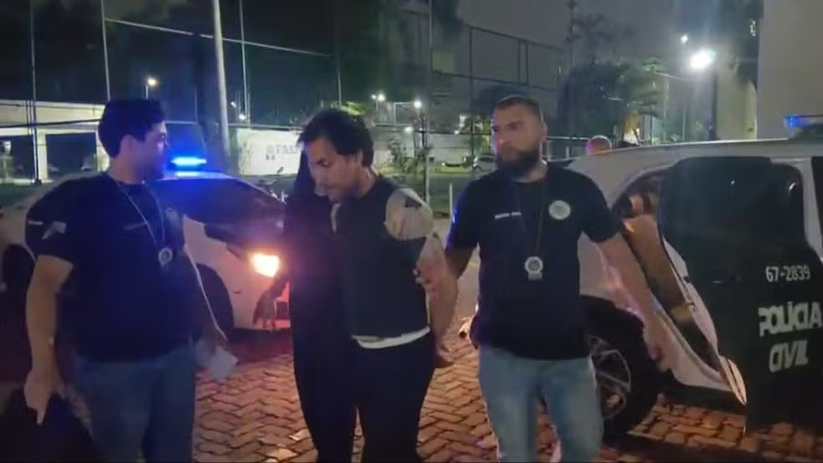 Dois policiais conduzem um homem próximo a uma viatura da Polícia Civil à noite