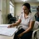 Mulher de camisa listrada sentada à mesa examinando documentos legais; 'PROCESSO' visível, placa 'ASSISTÊNCIA JURÍDICA'