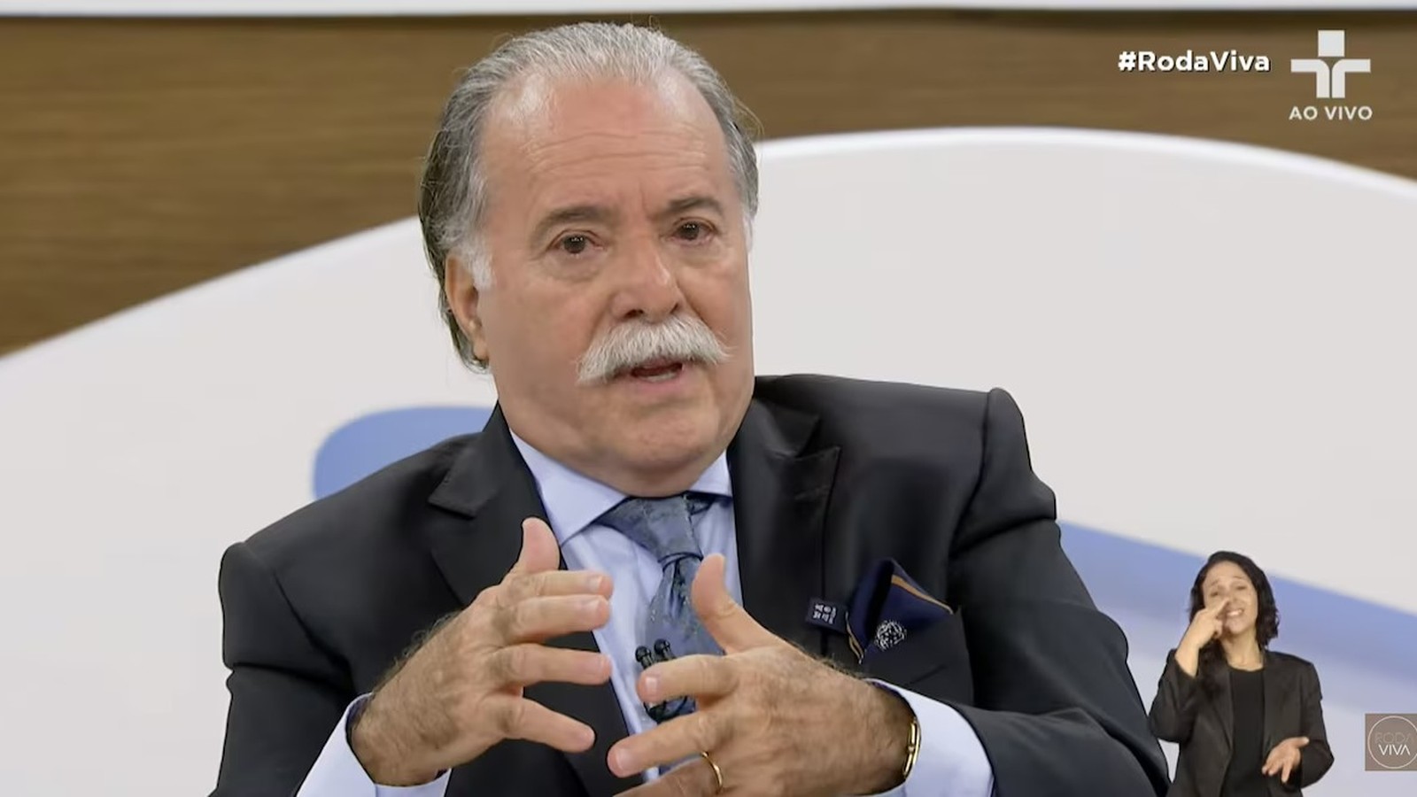 Tony Ramos em terno e gravata, gesticulando. Intérprete Libras e texto #RodaViva AO VIVO