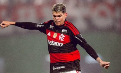 Jogador do Flamengo em campo, camisa preta e vermelha, cabelo claro e braços levantados