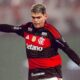 Jogador do Flamengo em campo, camisa preta e vermelha, cabelo claro e braços levantados