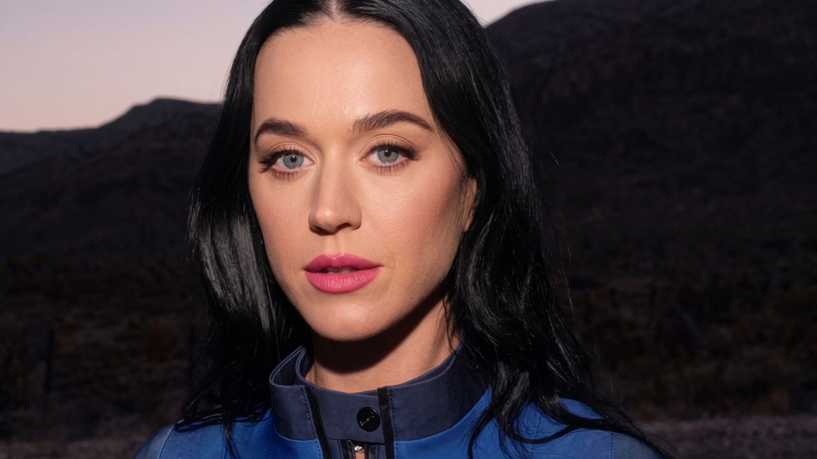 Katy Perry em close-up, cabelo escuro, olhos azuis, batom rosa e jaqueta azul