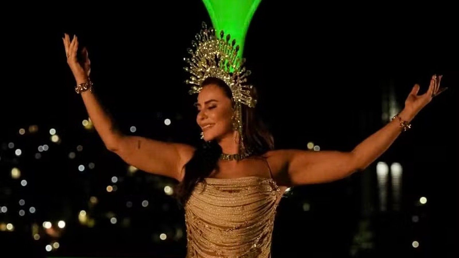 Paolla Oliveira sorrindo com coroa dourada e vestido brilhante, braços abertos contra fundo escuro.