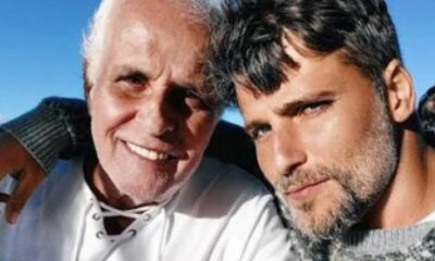 Dois homens sorrindo, um idoso de cabelo branco e um jovem barbudo com braço no ombro