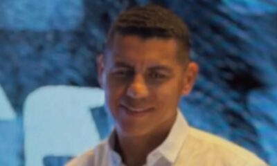 Homem com cabelo escuro e camisa branca, sorrindo levemente, contra um fundo azul desfocado