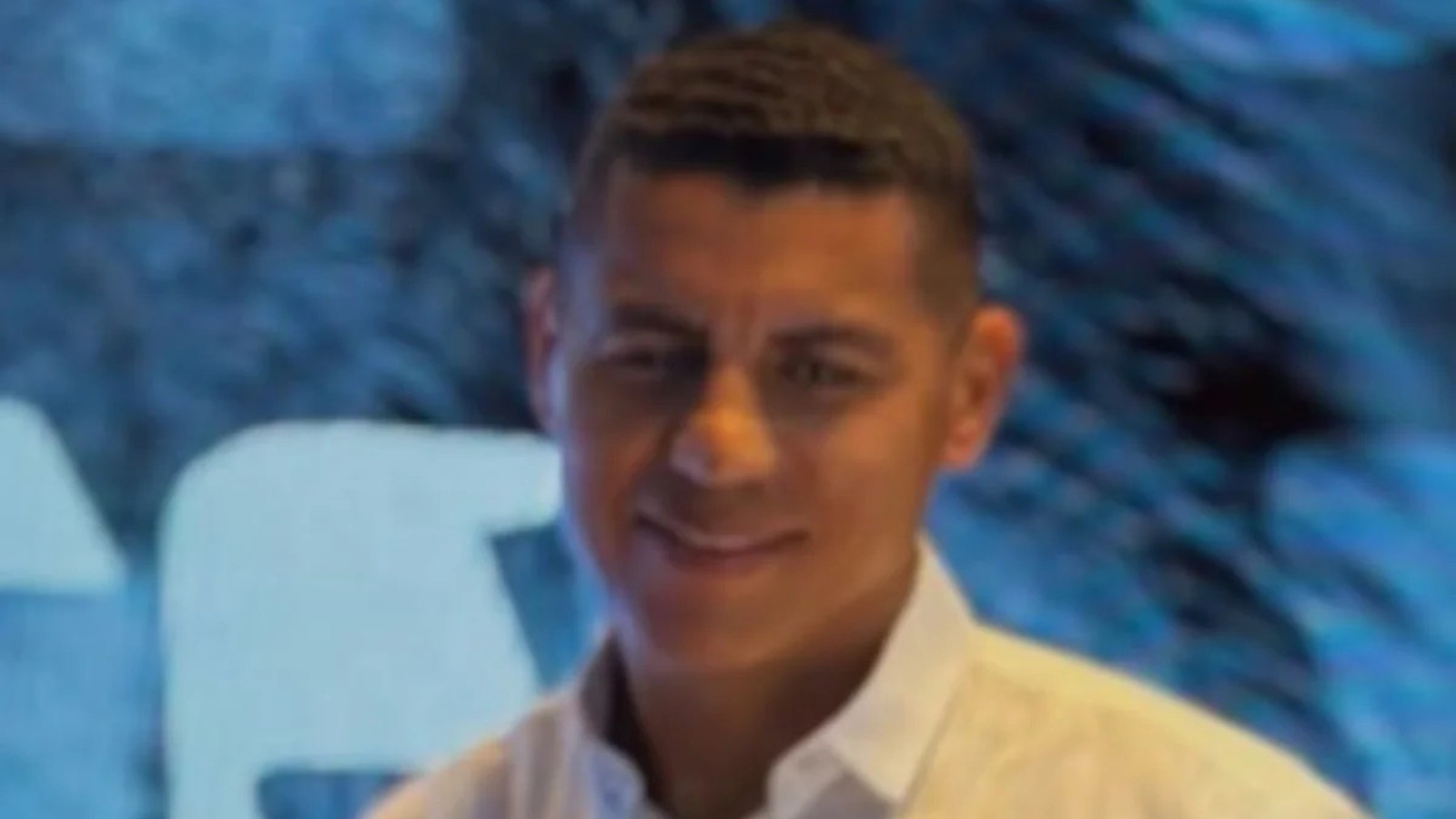 Homem com cabelo escuro e camisa branca, sorrindo levemente, contra um fundo azul desfocado